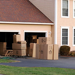 A tan house with white trim and brown shingles. In front of an open two-car garage are several groups of packs boxes sitting on the asphalt