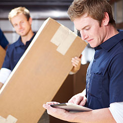Two males wearing matching navy polo shirts are shown, one maneuvering a long rectangular box and the other reading an inventory list. 