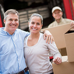 A middle-aged couple, a man with a blue collared shirt and a woman with a light brown long sleeve top, holding each other, smiling and posing in the foreground while out of focus behind them is a moving truck, some boxes and a mover holding a box.