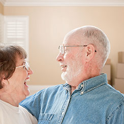 And elder couple, each wearing glasses, are facing each other and smiling, standing inside a residence with boxes behind them. 