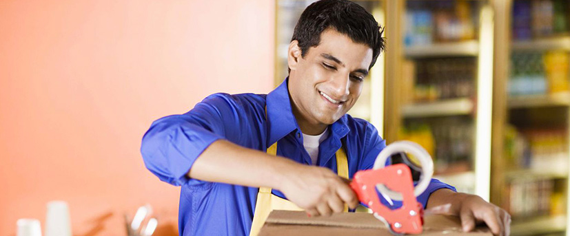 A man in a blue dress shirt and a yellow apron is taping a moving box with a hand-held tape dispenser.