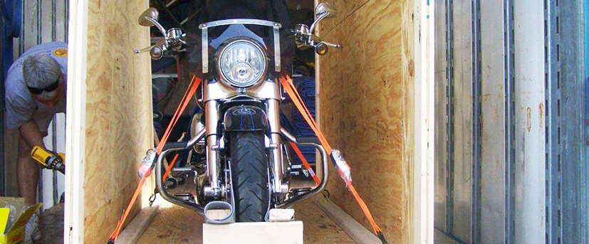 A motorcycle is seen strapped down and blocked within a plywood crate while a man with a drill continues to construct the crate around it.