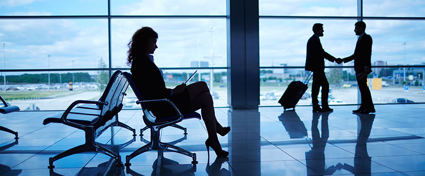 A blue- and black-hued image of an airport terminal with the dark silhouettes of three professionals are shown: A woman in a blazer and skirt seated in airport seating, working on her laptop in the foreground and two men in suits, shaking hands and one of them pulling his carry-on luggage.