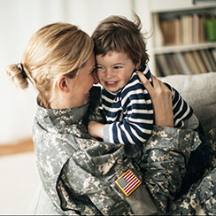 : A woman in military utility uniform is hugging her child who is wearing a blue and white striped long-sleeved shirt.