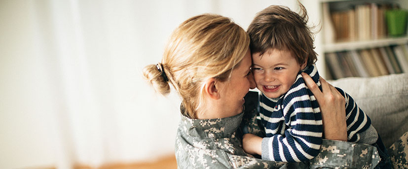 A young, female mother and service member is in her home wearing her utility uniform and holding her young toddler who is smiling.