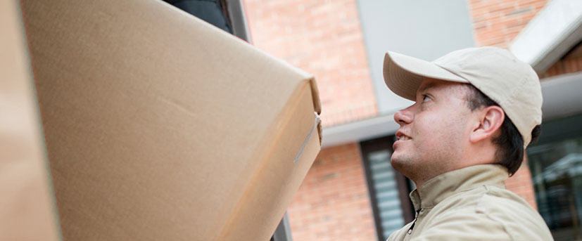 A male member of the moving company wearing a matching tan jacket and ball cap is looking up at another crew member as he a moving box is being lowered to him.