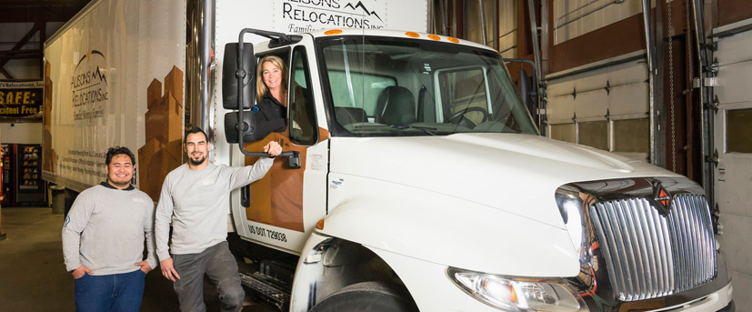 Owner of company, Alison, poses as she leans out of one of her moving truck cabs as two of her employees stand next to the truck smiling.